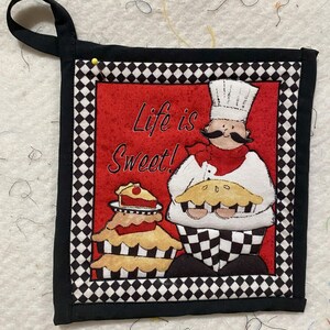 May include: A black and white checked pot holder with a red background and a cartoon chef holding a pie. The text "Life is Sweet!" is written in black on the red background.