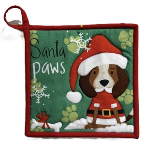 Puede incluir: Un salvamanteles navideño cuadrado y acolchado con borde rojo y lazo para colgar. El centro verde presenta un perro con traje y gorro de Papá Noel, con las palabras "Santa paws". Huellas de patas y copos de nieve decoran el fondo.