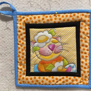 May include: A blue and orange quilted hot pad featuring a cat design. The cat is wearing a flower on its head and holding a flower in its paw. The background is yellow with white stripes.
