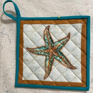 May include: A teal blue and brown quilted pot holder with a starfish design. The starfish is a light blue and brown color with intricate details.