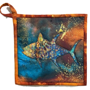 May include: Square, quilted potholder featuring a shark design. The shark is primarily blue and gold, set against a background of blue, orange, and brown. The potholder has a loop for hanging and a gold border.