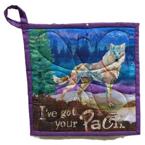 May include: Square quilted pot holder with a wolf design. The image features a wolf standing in front of a forest and mountains. The text "I've got your Pack" is printed on the bottom. The pot holder has a purple border and a hanging loop.
