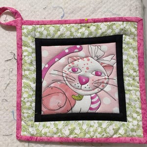 Può includere: Presa per pentole trapuntata rosa e verde con una cornice nera che presenta un gatto dei cartoni animati con un fiocco rosa. Il gatto tiene una palla rosa e ha pois rosa sul corpo.