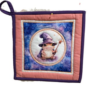 Può includere: Un sottopentola quadrato e trapuntato con un design stravagante. Il centro presenta una rana mago con una bacchetta a stella, su uno sfondo di galassia blu e viola. Il sottopentola ha un bordo rosa e viola e un anello per appenderlo viola.