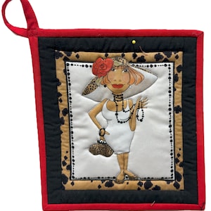 May include: A quilted potholder featuring a cartoon woman in a white dress, hat, and jewelry. The potholder has a red border, black and leopard print accents, and a red loop for hanging.