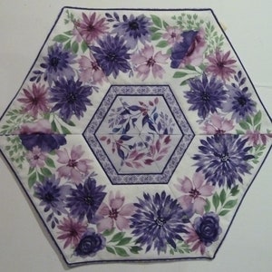 May include: A hexagonal fabric table runner with a white background and a floral pattern in shades of purple and pink. The flowers are arranged in a circular pattern around the center of the runner.