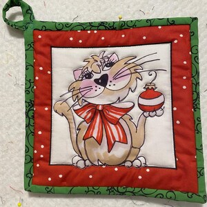 Könnte beinhalten: Ein roter und grüner gesteppter Topflappen mit weißem Hintergrund, auf dem ein Comic-Katze zu sehen ist, die einen roten und weißen Weihnachtsbaumschmuck hält. Die Katze trägt eine rot-weiß gestreifte Schleife.