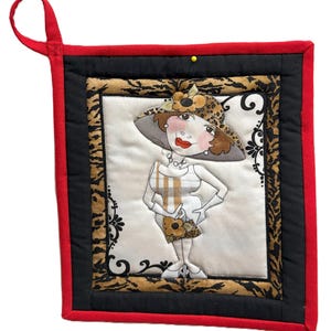 May include: Quilted hot pad featuring a cartoon woman in a leopard print hat and dress. The pad has a red border, black backing, and a leopard print and black frame around the central image. A red loop is attached for hanging.