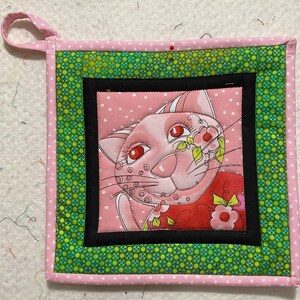 Peut inclure: Un dessous de plat carré en tissu matelassé avec un chat rose aux yeux rouges et des motifs floraux. Le chat est centré sur un fond rose à pois, encadré par un bord noir, un tissu vert à motifs et une bordure rose à pois. Une boucle rose est attachée pour suspendre.