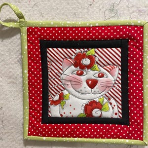 Puede incluir: Un posavasos acolchado rojo y blanco con un borde verde. El centro presenta un gato blanco con flores rojas y un fondo a rayas rojo y blanco.