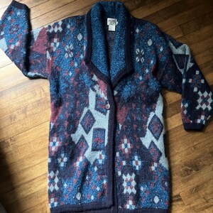May include: A long, button-down cardigan in shades of blue, burgundy, and gray with a geometric pattern. The sweater has a shawl collar and long sleeves. The garment is made of a textured knit fabric.