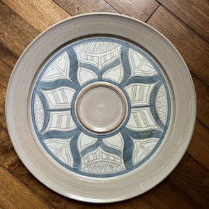 May include: A handcrafted ceramic serving platter with a beige rim and a central design in white and blue. The center features a small, recessed area. The platter is signed "KRAUSE '94".