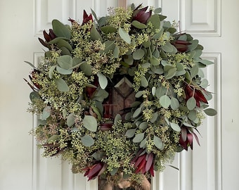 May include: A wreath made of eucalyptus and burgundy flowers hangs on a white door.