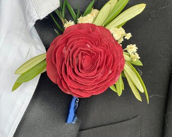 May include: A red rose boutonniere with green leaves and white flowers. The boutonniere is pinned to a black suit jacket.