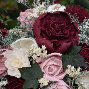 May include: A bouquet of burgundy, blush pink, and white wooden roses with greenery and white baby's breath.