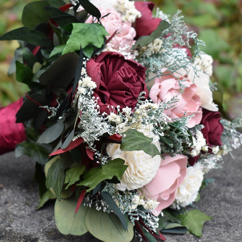 May include: A bridal bouquet with burgundy and blush pink silk flowers, greenery, and white baby's breath.
