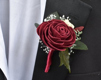 May include: A burgundy rose boutonniere with white baby's breath and green leaves, perfect for a wedding or special occasion.