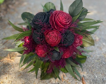May include: A bouquet of burgundy and black wooden roses with greenery. The bouquet is arranged in a circular shape and is sitting on a gray surface.