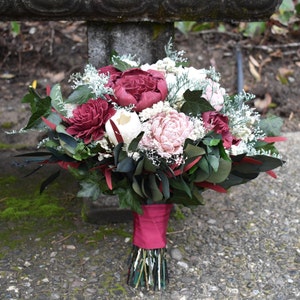 May include: A bouquet of burgundy and blush silk flowers with greenery and white accents. The bouquet is tied with a burgundy ribbon.