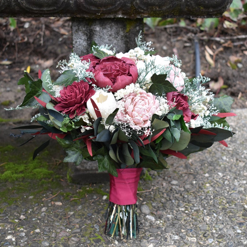 May include: A bouquet of burgundy and blush silk flowers with greenery and white accents. The bouquet is tied with a burgundy ribbon.