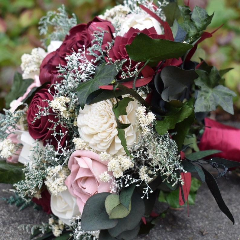 May include: A bridal bouquet with burgundy, blush pink, and white flowers. The bouquet is made of wood and features greenery.