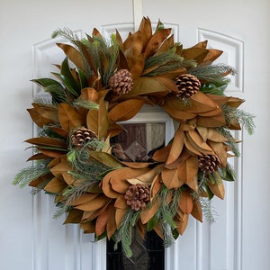 May include: A brown magnolia leaf wreath with pine sprigs and pine cones. The wreath is hanging on a white door.