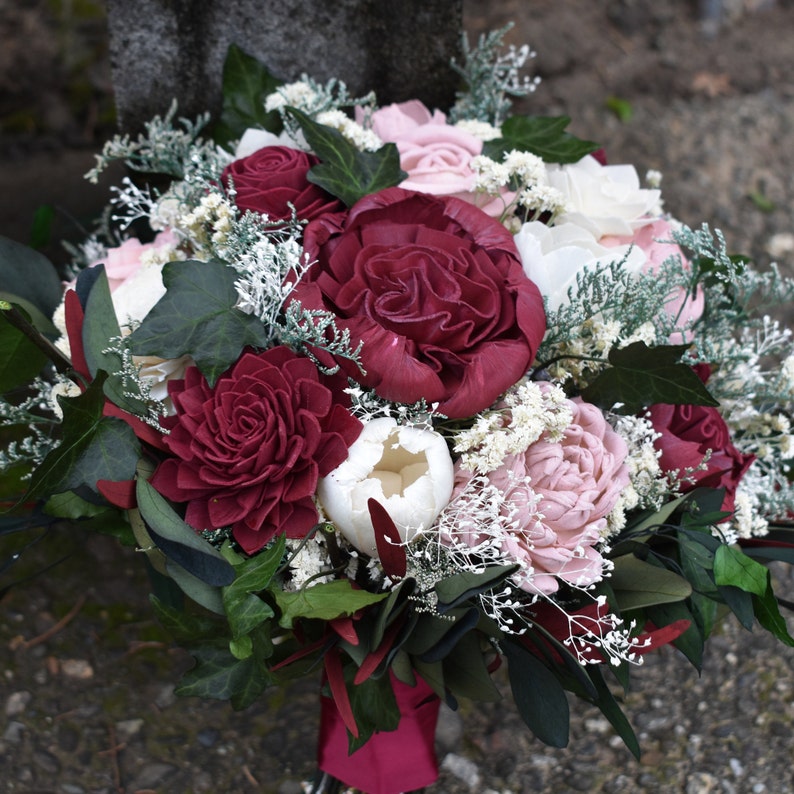 May include: A bouquet of burgundy and blush-colored silk flowers with white accents. The bouquet is tied with a burgundy ribbon.