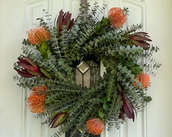 May include: A circular wreath featuring green eucalyptus leaves and vibrant orange pincushion protea flowers, with accents of burgundy protea. The wreath is displayed on a white door, creating a welcoming and natural aesthetic.