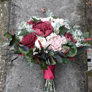 May include: A bouquet of burgundy and blush silk flowers with white and green foliage. The bouquet is tied with a burgundy ribbon.