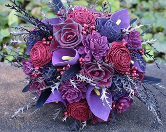 May include: A bouquet of burgundy and purple silk flowers with black stems and leaves. The bouquet is arranged in a circular shape and features a variety of flower types, including roses, calla lilies, and carnations.