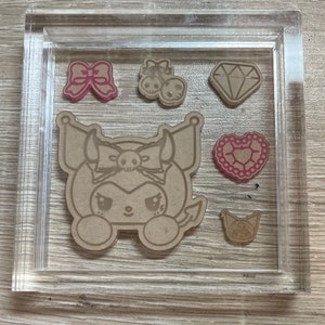 Puede incluir: Un juego de sellos de acrílico transparente con seis diseños diferentes. Los diseños incluyen un corazón, un diamante, un lazo, una calavera con cerezas, un gato y un personaje con cuernos y un lazo. Los sellos están sobre un fondo marrón.