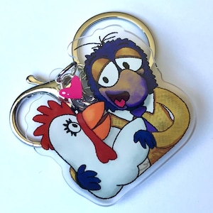 Può includere: Un portachiavi in acrilico trasparente con un personaggio dei cartoni animati viola e marrone che tiene un pollo bianco con una cresta rossa. Il portachiavi ha un anello di metallo argentato.
