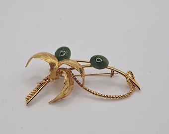 May include: A gold-tone brooch featuring a stylized palm tree with two green jade stones. The brooch has a twisted rope design.