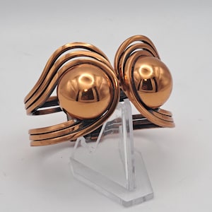 May include: A pair of copper-colored cuff bracelets. Each bracelet features a large, polished sphere at the center, framed by curved, layered bands. The design is symmetrical and geometric, with a reflective surface. The bracelets are displayed on a clear stand.