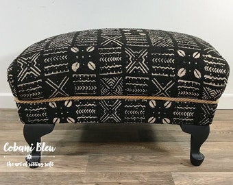 May include: Black and white patterned ottoman with a tan rope trim and black wooden legs. The ottoman is sitting on a wood floor.