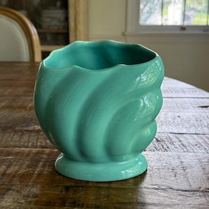 May include: A turquoise ceramic vase with a swirled design. The vase has a round base and a wide opening. The vase is sitting on a wooden table. The vase is a decorative item.
