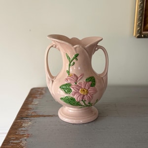 May include: A pink ceramic vase with two handles and a flared top. The vase features embossed pink flowers with yellow centers and green leaves. The vase is on a pedestal base and is likely vintage.