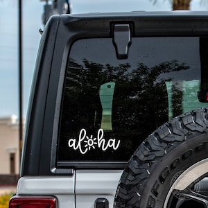 May include: White vinyl decal with the word "Aloha" and a sun graphic on a grey car window.