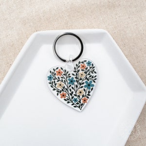 May include: A heart-shaped keychain with a floral design. The clear acrylic keychain features a pattern of blue, orange, and yellow flowers with black stems. A silver metal ring is attached at the top.