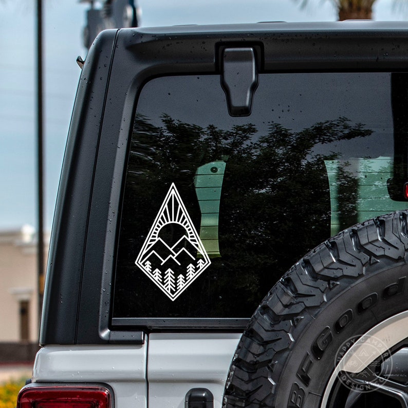 May include: White vinyl decal of a mountain range with a sun rising behind it, inside a diamond shape. The decal is on the back window of a silver car.