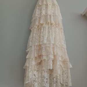 Puede incluir: Una falda larga de encaje de color marfil con múltiples capas de volantes. La falda está hecha de encaje floral delicado y tiene una silueta suave y fluida.
