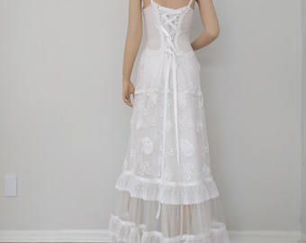May include: White lace wedding dress with a fitted bodice and a flowing skirt. The dress features a lace-up back and multiple layers of ruffles.
