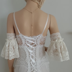 May include: White lace wedding dress with a lace bodice and a lace overlay skirt. The dress has a low back with a white satin ribbon tie and a delicate gold chain necklace.
