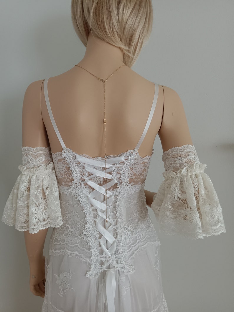 May include: White lace wedding dress with a lace bodice and a lace overlay skirt. The dress has a low back with a white satin ribbon tie and a delicate gold chain necklace.