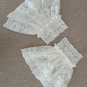 May include: Two white lace cuffs with a delicate floral pattern. The cuffs are made of a soft, sheer fabric and have a ruffled edge.