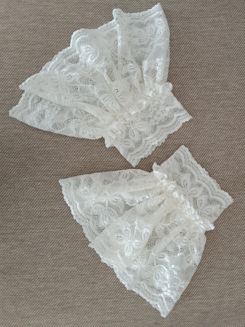 May include: Two white lace cuffs with a delicate floral pattern. The cuffs are made of a soft, sheer fabric and have a ruffled edge.