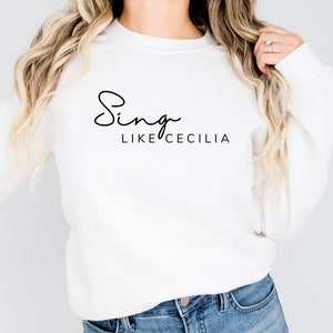 May include: White long-sleeve sweatshirt with black text that reads "Sing like Cecilia".