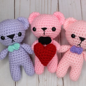Può includere: Tre orsacchiotti di peluche all'uncinetto, uno viola, uno rosa e uno rosa chiaro, con occhi neri e un naso nero. L'orso centrale tiene un cuore rosso all'uncinetto.
