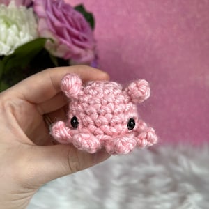 May include: A small pink crocheted octopus with black eyes. The octopus is being held in a hand.
