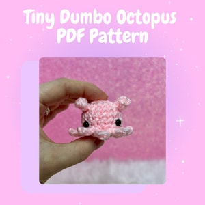 Könnte beinhalten: Ein kleiner rosa gehäkelter Dumbo-Oktopus mit schwarzen Augen. Der Oktopus wird von einer Hand vor einem rosa Hintergrund gehalten. Der Text "Tiny Dumbo Octopus PDF Pattern" befindet sich oben im Bild.